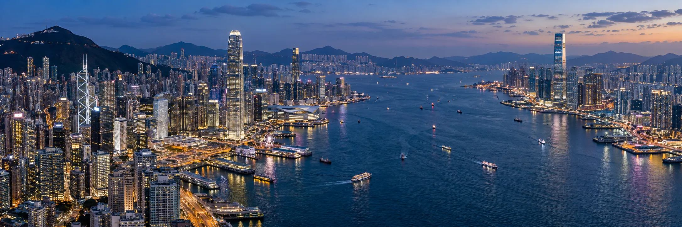 Victoria Harbour at dusk, with lit towers on both shores and ferries crossing the water.