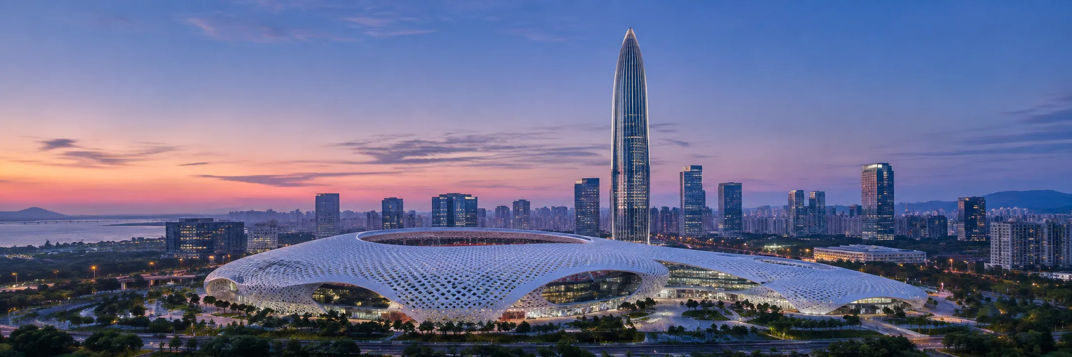 Nanshan, Shenzhen at dusk, with a white lattice stadium in front, a lit glass tower behind it, and the bay beyond.